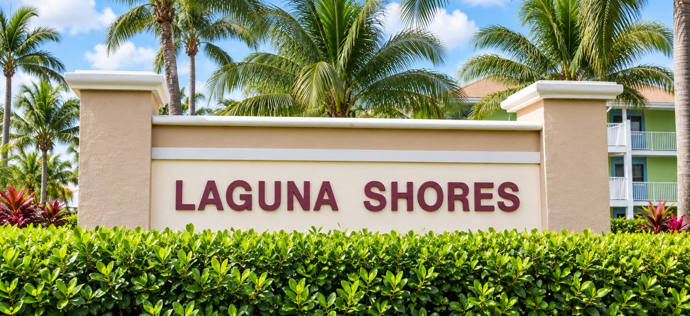 The Laguna Shores entrance sign on Fort Myers Beach, Florida: deep red lettering on a low stucco wall between two capped pillars, with a clipped hedge in front, royal palms behind and a balconied building beyond on the right.