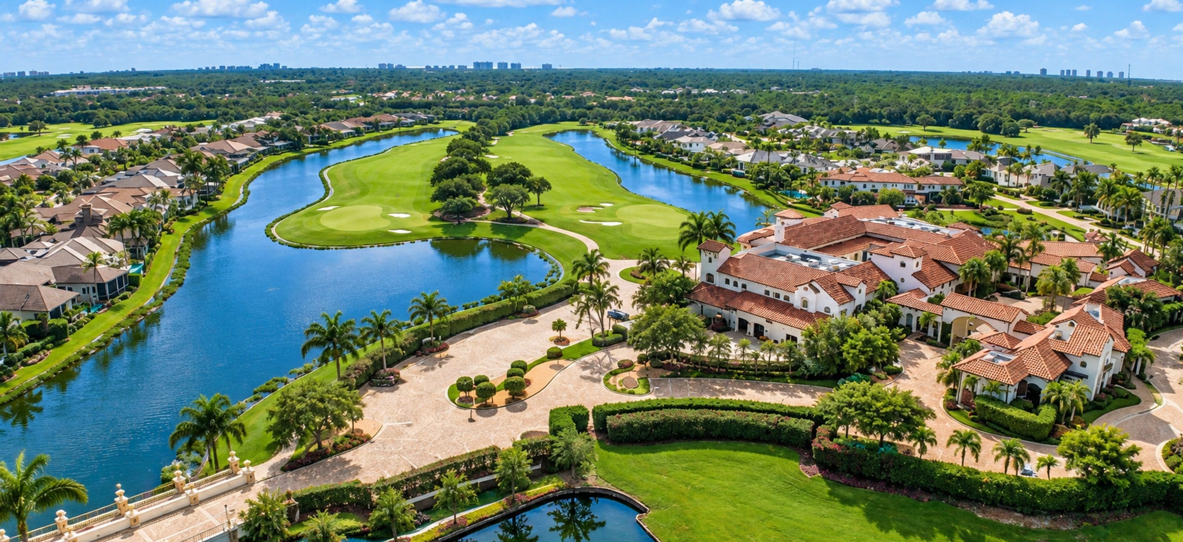 An aerial view of Talis Park in Naples, Florida: the Vyne House clubhouse with its terracotta roofs beside the golf course, fairway and greens running between two lakes, homes along the water and a balustraded bridge in the foreground.